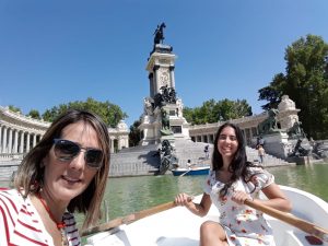 Carla and her mom, Nelly are on a row boat on a lake at the Parque del Retiro en Madrid, Spain. Carla wears a white dress with orange flowers and holds the oars. Nelly takes the picture selfie-style and wears a red and white striped shirt, a red necklace, and sunglasses. Behind them is the Monument to Alfonso XII. The sky is clear and blue.