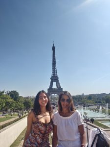 Carla and her mom, Nelly, sit with the Eiffel Tower seen behind them. Carla wears a floral dress and Nelly wears a white top with white pants. The sky is blue and cloudless.