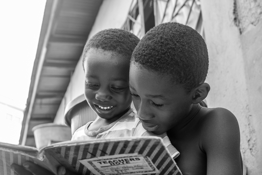 Black and white photo by Samuel Aye-Gboyin featuring an older boy reading with a smiling younger boy