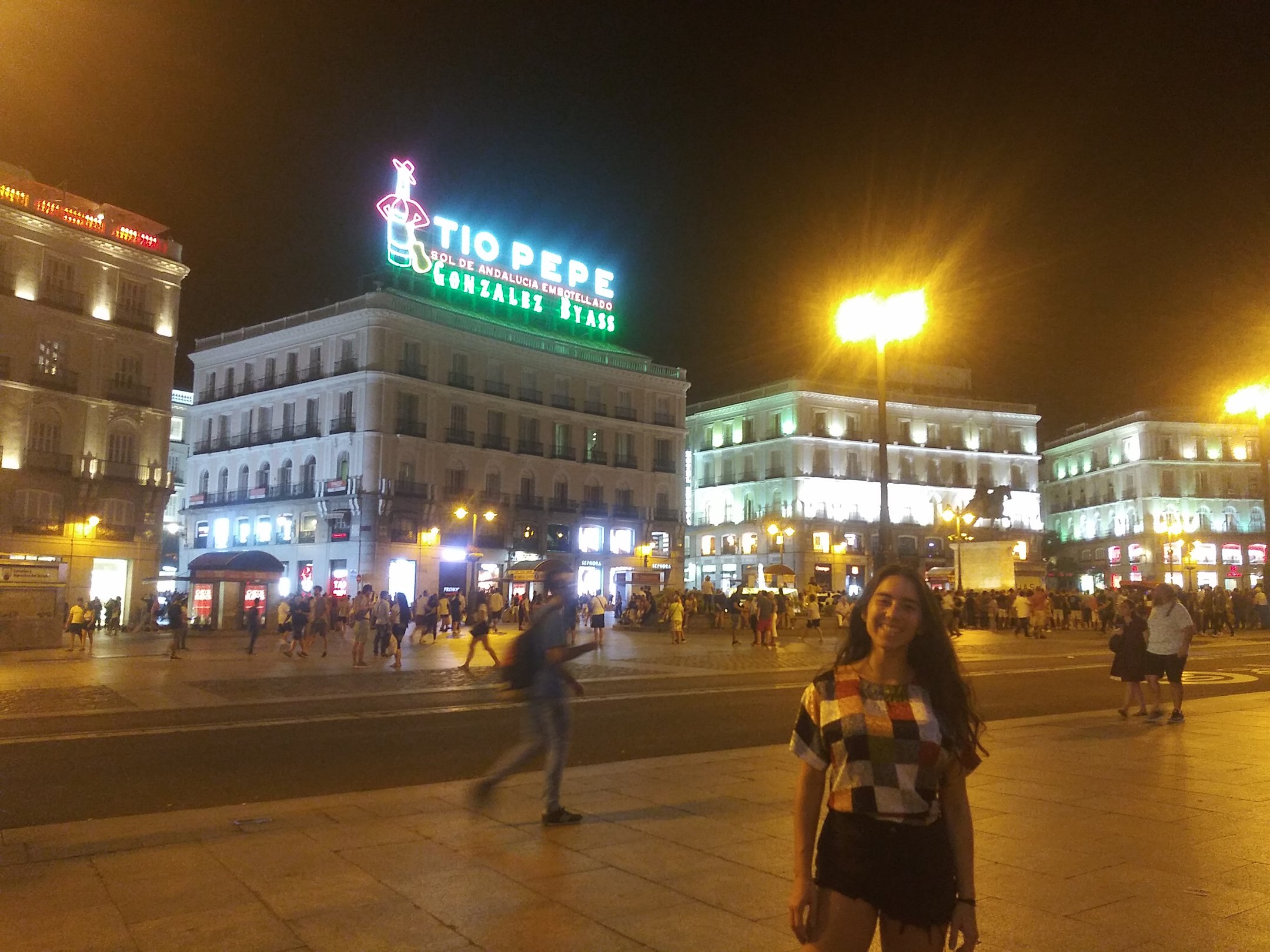 Carla wears a patched shirt with different patterns and black shorts. She stands in front of Puerta del Sol in Madrid, Spain at night where a bright "Tio Pepe" neon sign shines behind her. The plaza is bustling with people.