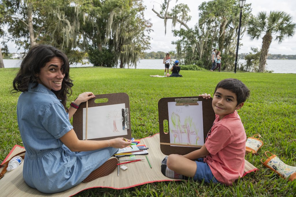 Mother and son show their landscape drawings while sitting on a map on a lawn by a lake