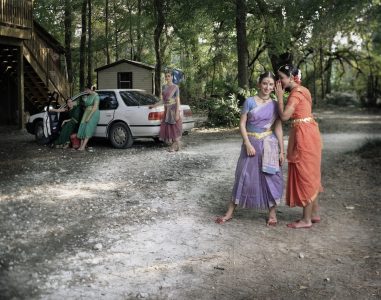 On a dirt road two women whisper in the foreground while more people stand by and lean on a white car in the background