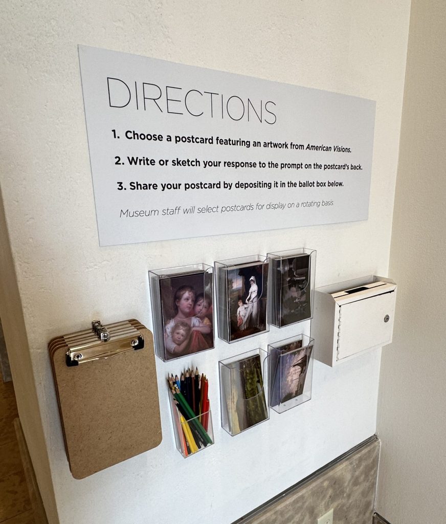 Postcards featuring reproductions of artworks, clipboards, and colored pencils hang next to a ballot box on a gallery wall under a sign that says "Directions and gives instructions for how to use the cards.