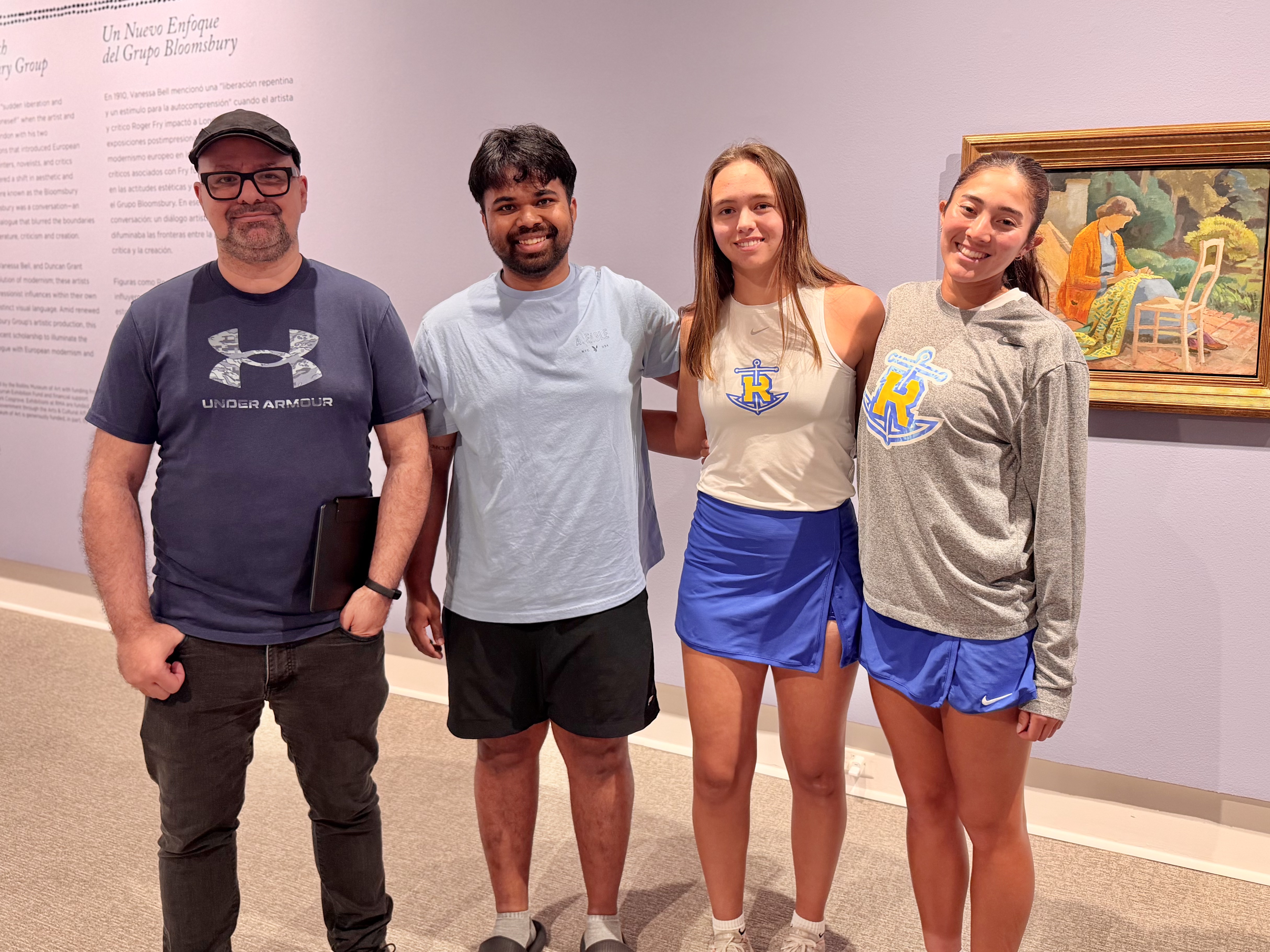Photograph of four people standing side by side in Rollins Museum of Art gallery setting with artwork and text on a purple wall behind them. Two individuals wear matching athletic outfits with blue shorts and logo shirts, while the other two wear casual clothing, highlighting a casual group visit or event.